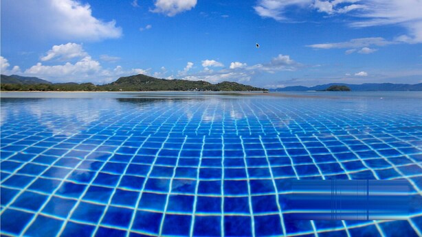 Una piscina azul cristalina con un diseño de cuadrícula, rodeada por un paisaje natural con colinas y un cielo despejado.