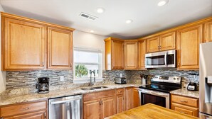 A modern kitchen with wooden cabinets, granite countertops, stainless steel appliances, and a window with a view of palm trees. - East End Escape | Ocean Views, Spacious Deck & Quiet Beach Area Near Public Access (Folly Beach)