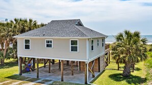 A two-story house elevated on stilts with a balcony, surrounded by palm trees and a view of the ocean. - East End Escape | Ocean Views, Spacious Deck & Quiet Beach Area Near Public Access (Folly Beach)