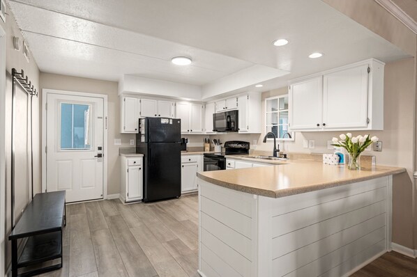 A modern kitchen with white cabinetry, a black refrigerator, a microwave, and a sink. - First Light: PierView Oceanfront Condo, Contemporary Vibes & Stunning Views (Nags Head)