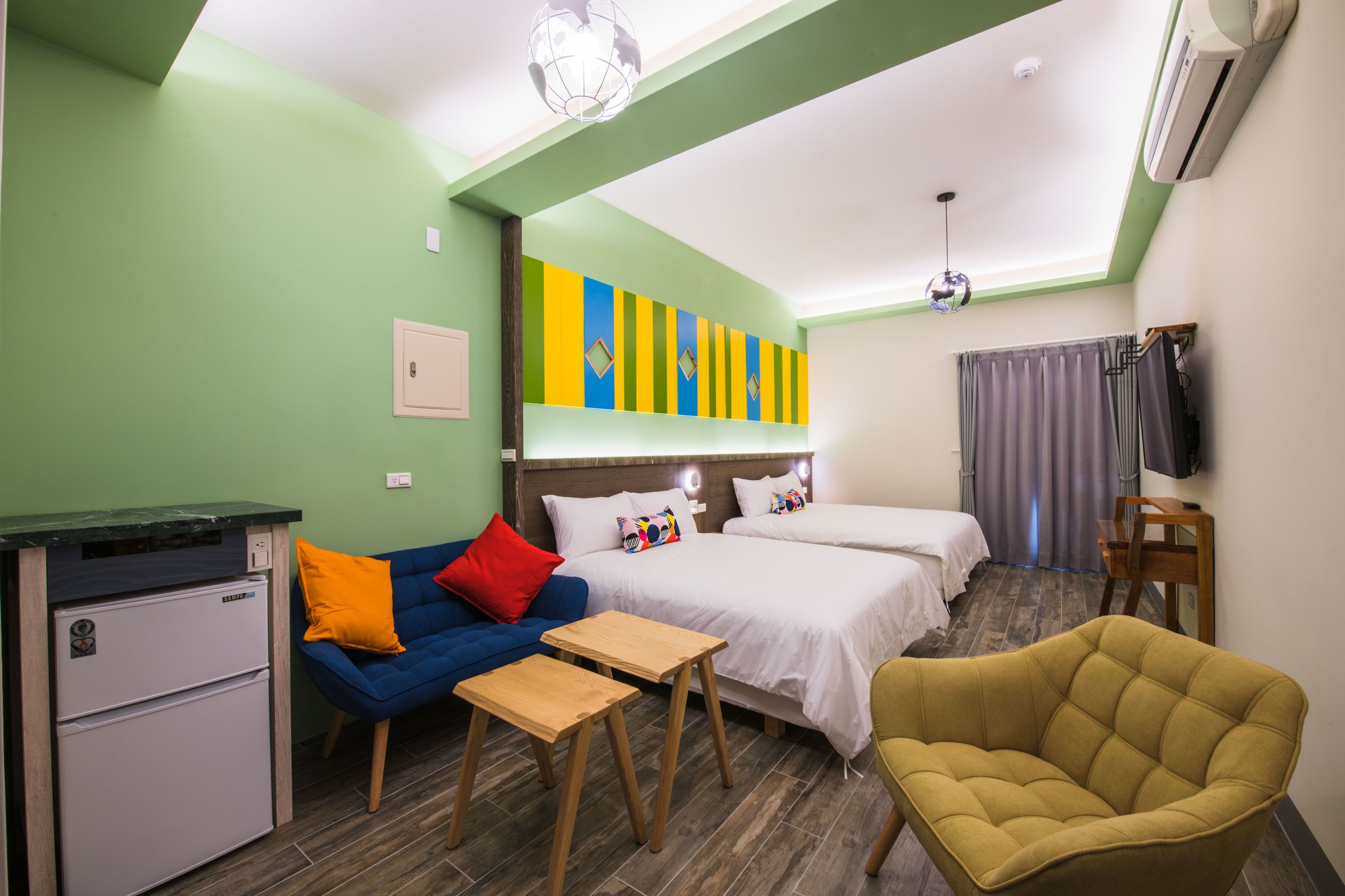 Une chambre d’hôtel équipée d’une kitchenette, d’un canapé, d’un fauteuil jaune, de deux lits, d’une télévision et d’une décoration murale colorée.