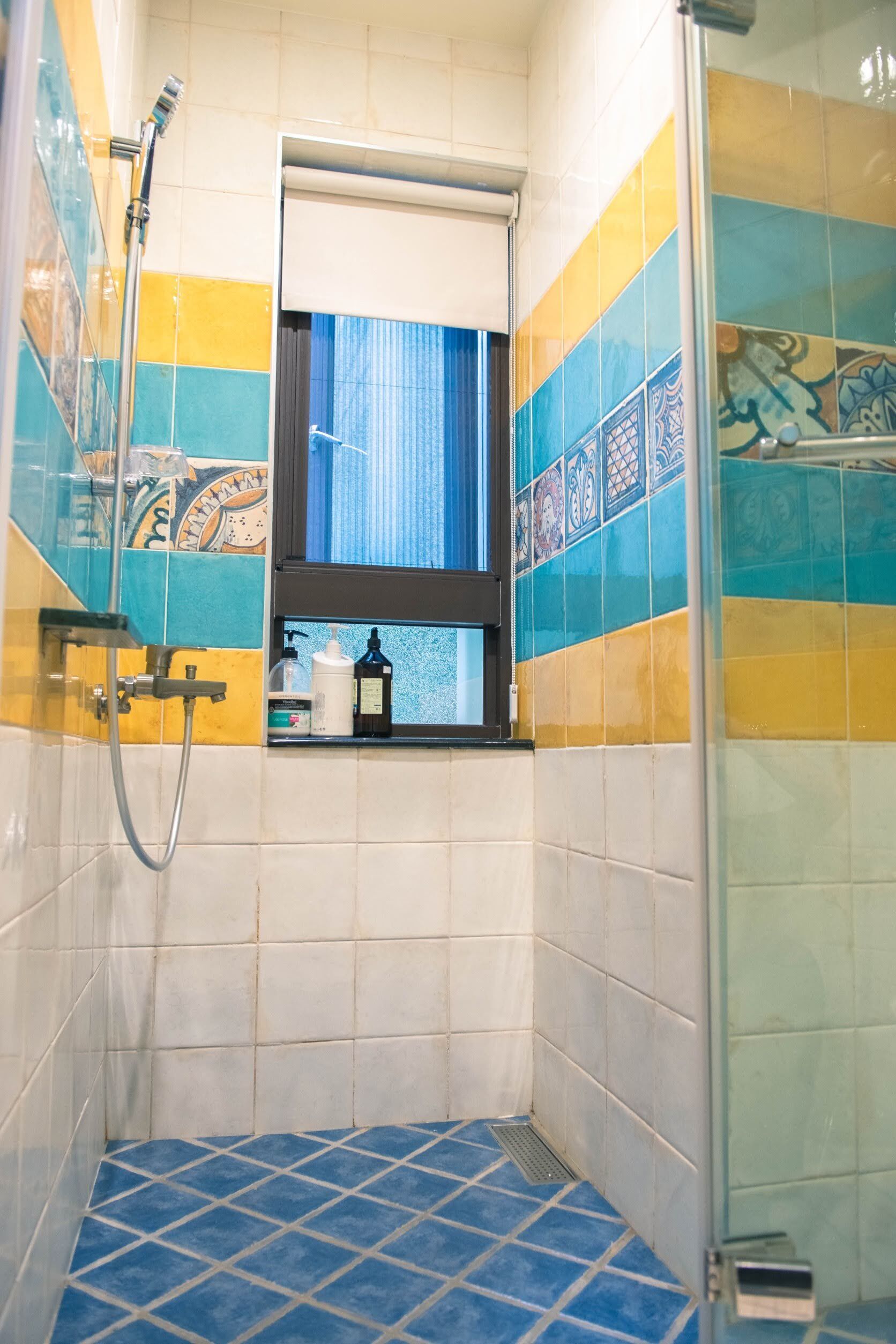 Ein kleines Badezimmer mit blau-gelben Fliesen, einer Dusche und einem Fenster mit Vorhang.