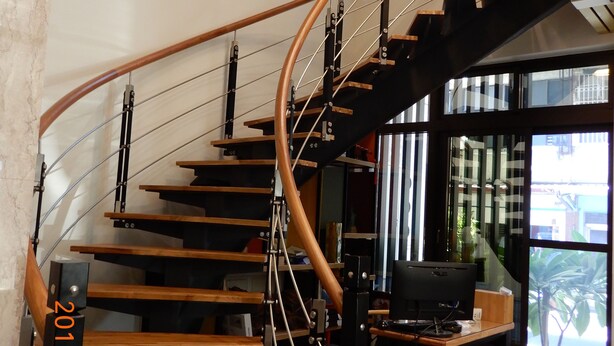 Una escalera espiral moderna con escalones de madera y barandillas de metal, que conduce a una zona del segundo piso con un escritorio y un ventanal grande.