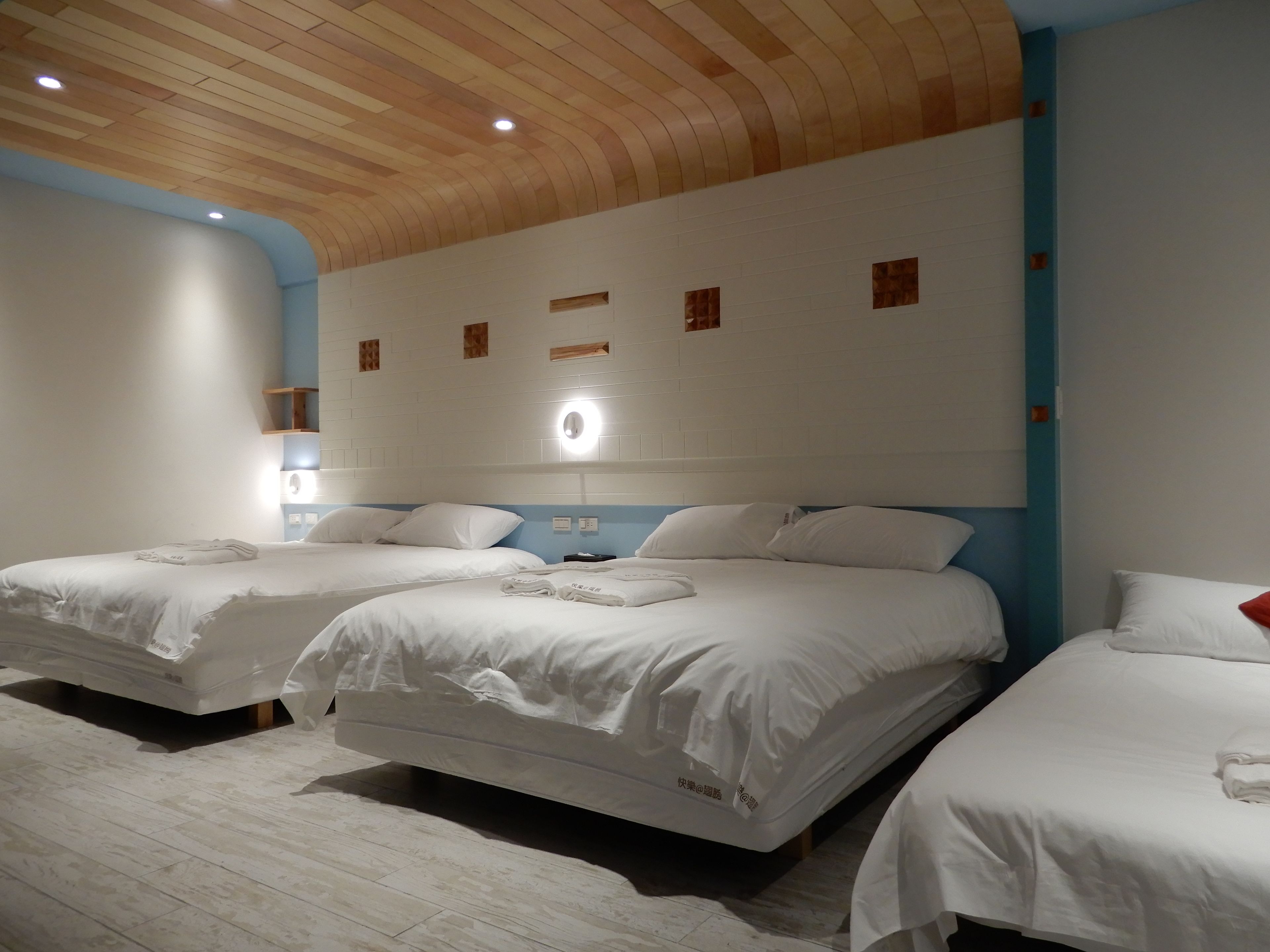 Une chambre d’hôtel avec trois lits, un plafond en bois, des murs blancs et un mur d’accent bleu.