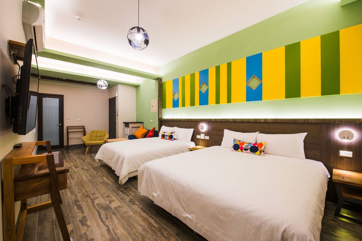 A hotel room with two beds, a desk, a chair, a TV, and a colorful wall decoration.