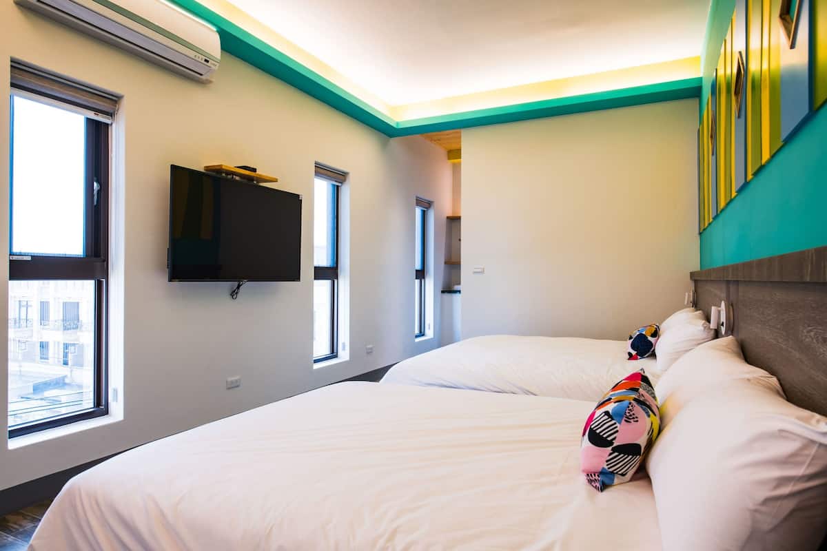 A hotel room with two beds, a TV mounted on the wall, and large windows.
