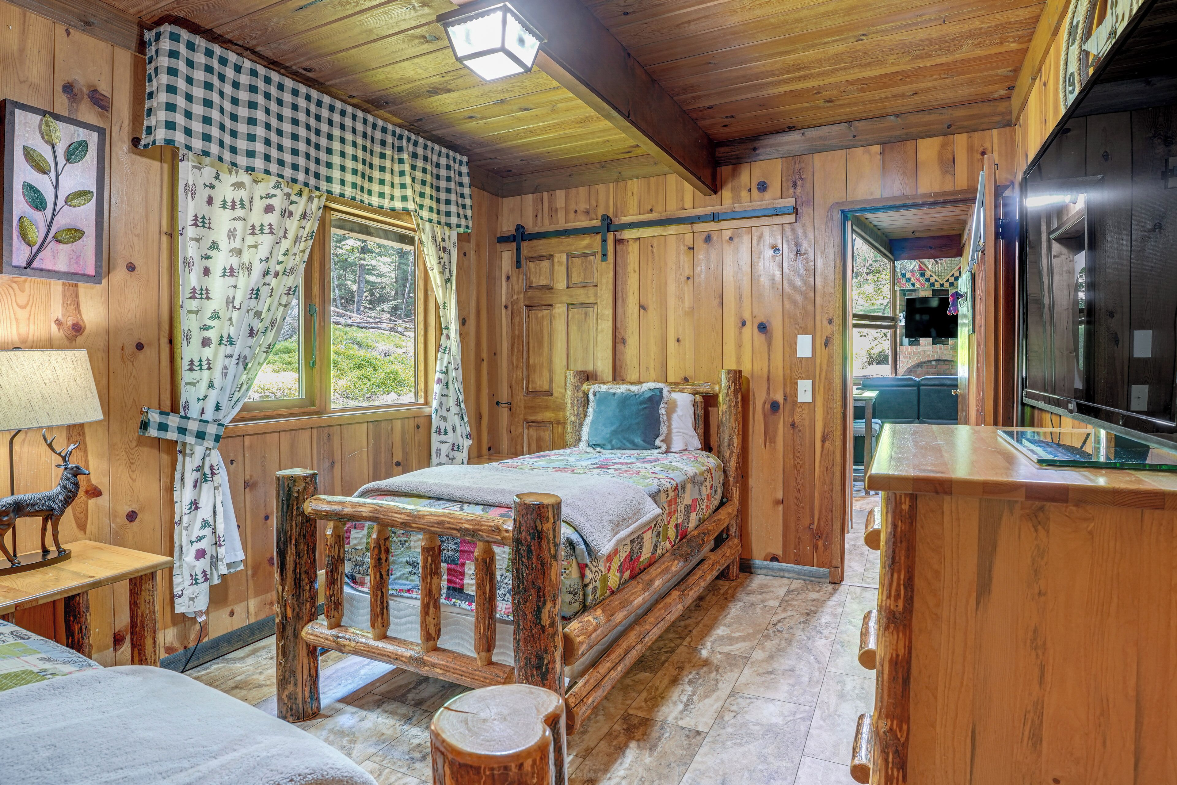 Interno di una baita in legno, con un letto, una panchina di legno, un tavolo e una finestra con tende.