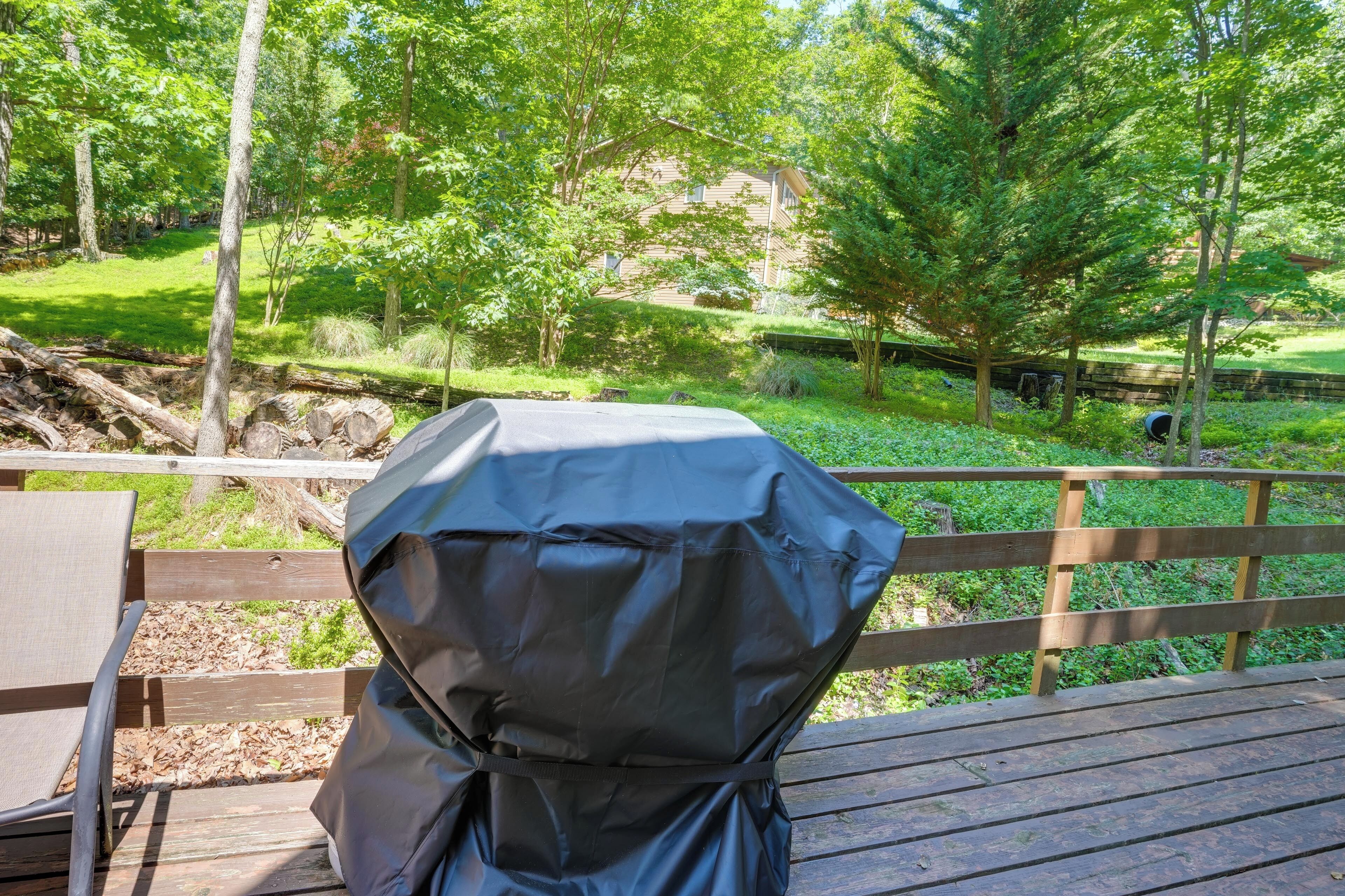 A wooden deck with a barbecue grill, surrounded by lush greenery and a distant building.