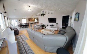Amplia sala de estar con sofá gris, una mesa de centro y televisión. Se observa un área de cocina al fondo con una mesa y sillas de comedor.