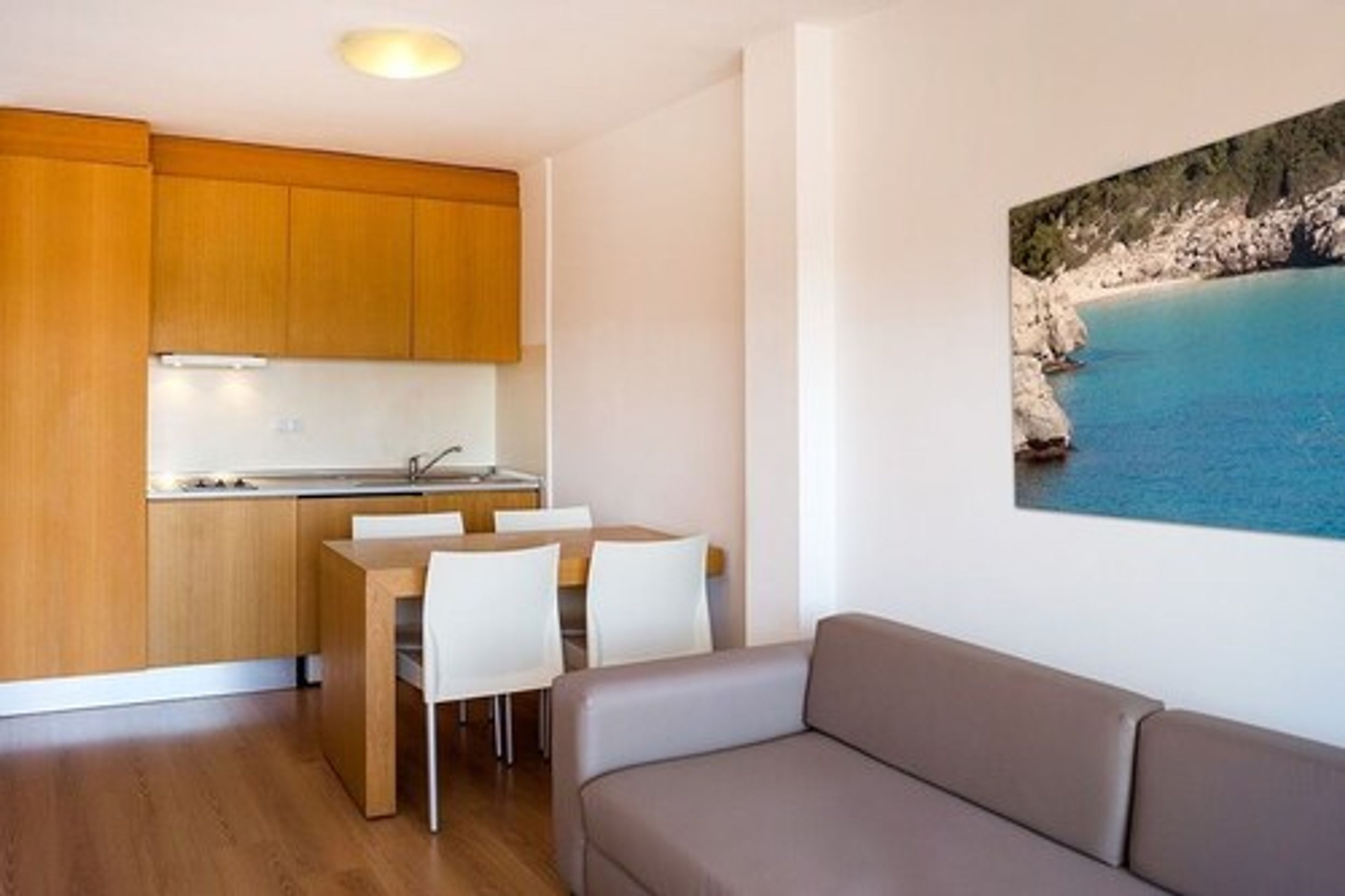 Una cocina moderna con armarios de madera, un comedor con sillas blancas y un sofá beige. En la pared, un cuadro grande de un paisaje costero.