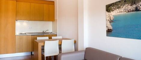 Una cocina moderna con armarios de madera, un comedor con sillas blancas y un sofá beige. En la pared, un cuadro grande de un paisaje costero.