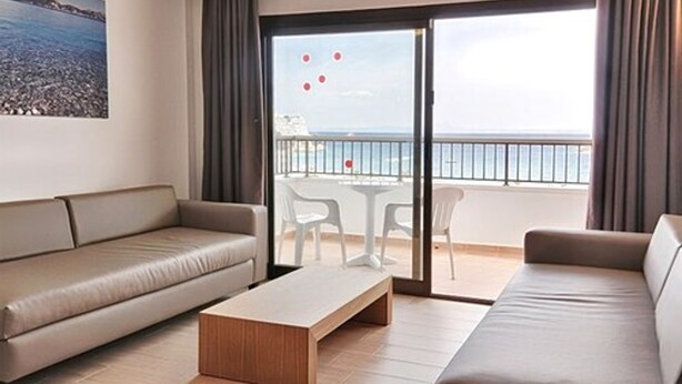 Una camera d'hotel moderna con un divano color beige, un tavolino in legno e un balcone con vista sul mare.