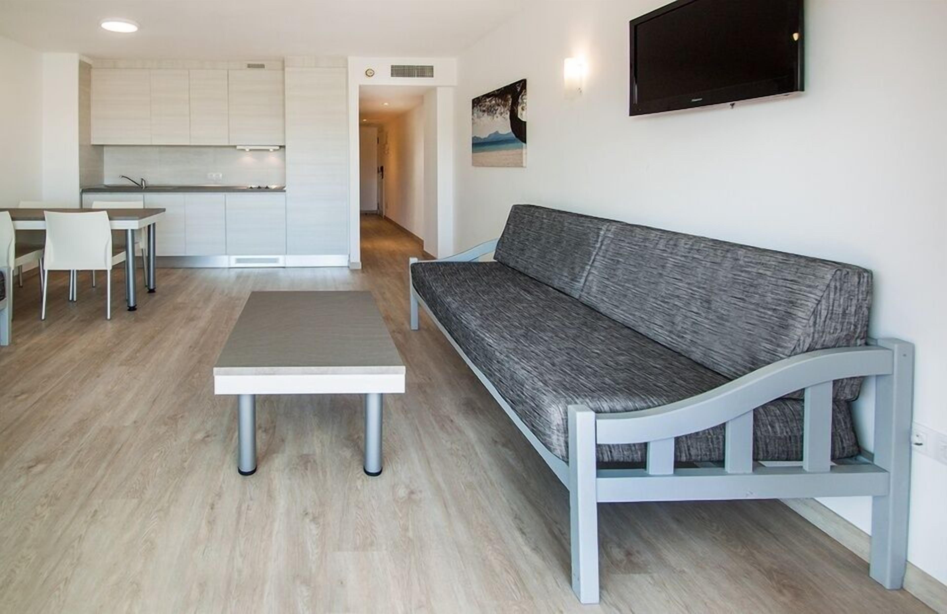 A modern living room with a grey sofa, a white coffee table, and a dining area with a white table and chairs. A kitchen with white cabinets is visible in the background.