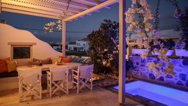 An outdoor seating area with a table and chairs, a covered pergola, and a view of the cityscape at night.