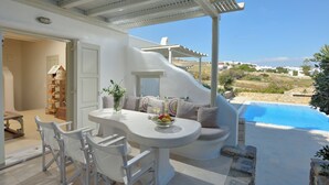A modern outdoor dining area with a white table and chairs, a bowl of fruit, and a view of a pool and landscape.