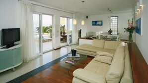 Ein modernes Wohnzimmer mit einer Couch, einem Couchtisch und einem Fernseher. Eine Glastür öffnet sich zu einem Balkon mit einem Pool.