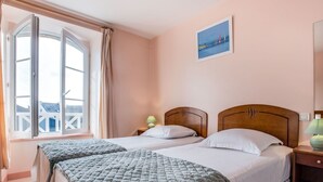Una camera d'albergo con due letti, un'ampia finestra con tende, un quadro appeso al muro e un comodino con una lampada.