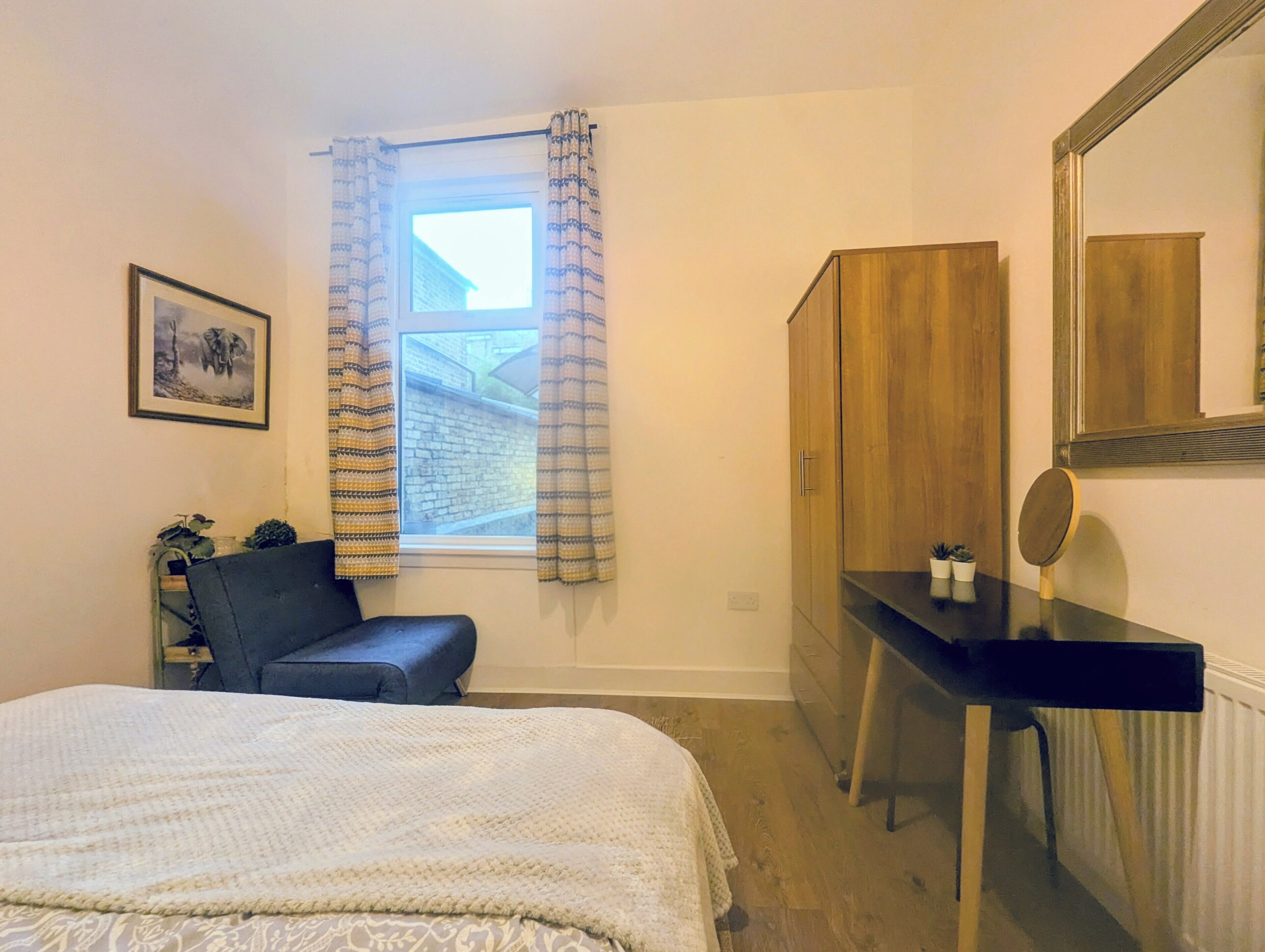 A bedroom with a bed, a desk, a mirror, a window with curtains, a framed picture, and a radiator.