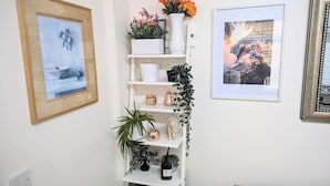 Pared blanca con tres cuadros enmarcados, estante blanco con adornos y una mesa con mantel a cuadros.