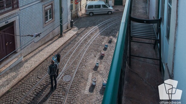 A balcony overlooking a cobblestone street with a tram track, buildings with balconies, and a distant view of the sea.
