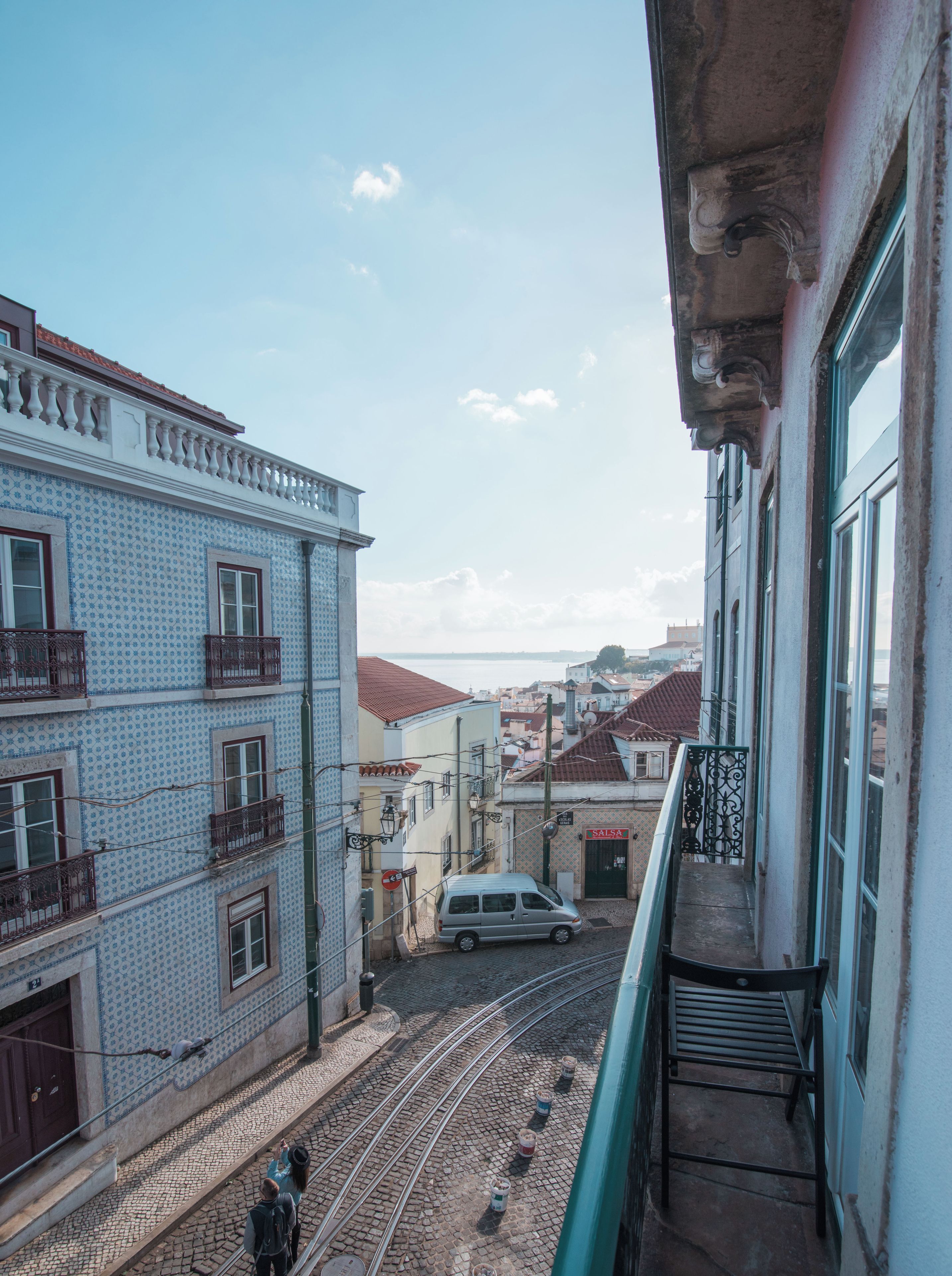 Un balcón con vistas a una calle empedrada con una vía de tranvía, edificios con balcones y una vista distante del mar.