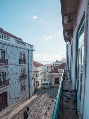 Uma varanda com vista para uma rua de calçamento, um trilho de bonde, edifícios com varandas e o mar ao longe.