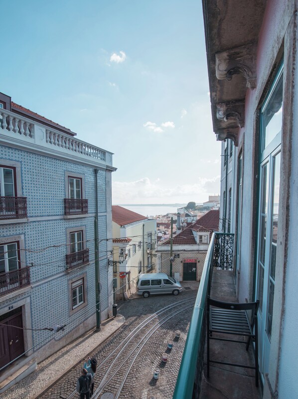 Un balcon donnant sur une rue pavée avec une voie de tramway, des immeubles dotés de balcons et une vue lointaine sur la mer.