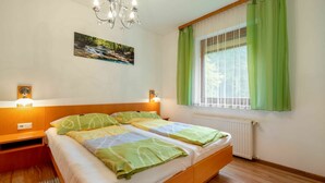 Ein Doppelbett mit grün-weißer Bettwäsche, ein Holz-Kopfteil, eine Chandelüster, ein Bild einer Wasserfall und ein Fenster mit grünen Vorhängen sowie ein Heizkörper.