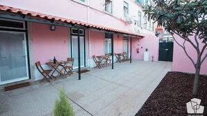 A pink building with a tiled roof, outdoor seating area with wooden chairs and tables, and a small tree in the foreground.
