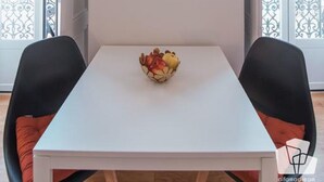 A dining area with a white table, two black chairs, and a bowl of fruit. There are three decorative plates on the wall.