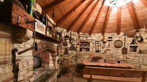 Una habitación rústica con paredes de piedra, vigas de madera, un horno de ladrillo, estantes con diversos objetos y una mesa de madera.