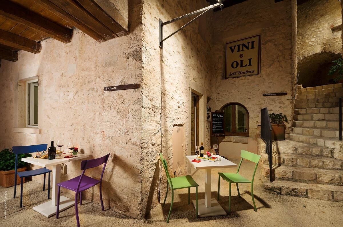 Ein Essbereich im Freien mit rustikalen Steinmauern, einem Schild mit der Aufschrift "VINI e OLI dell'Umbria" und einem Tisch, der mit Weingläsern und einer Flasche gedeckt ist.