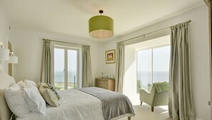 A bedroom with a bed, a nightstand, a dresser, a chair, and a large window with curtains. - Stunning coastal home with panoramic sea views in Porlock Weir - 6 bed / 6 bath (Minehead)