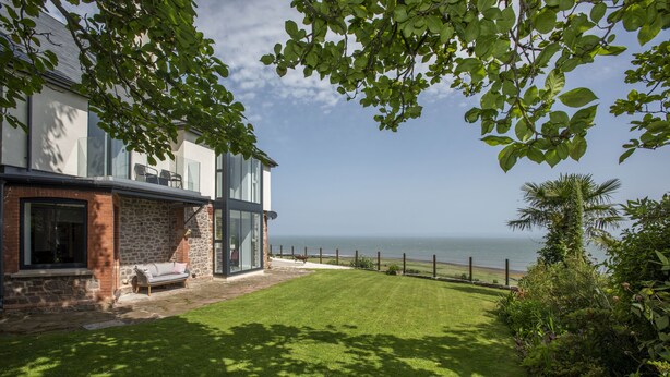A modern house with a stone facade, a glass conservatory, and a well-maintained lawn leading to a coastal view. - Stunning coastal home with panoramic sea views in Porlock Weir - 6 bed / 6 bath (Minehead)