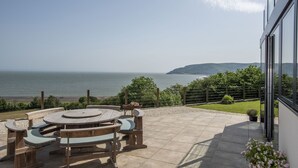 A patio with a wooden table and benches overlooking a body of water and hills. - Stunning coastal home with panoramic sea views in Porlock Weir - 6 bed / 6 bath (Minehead)