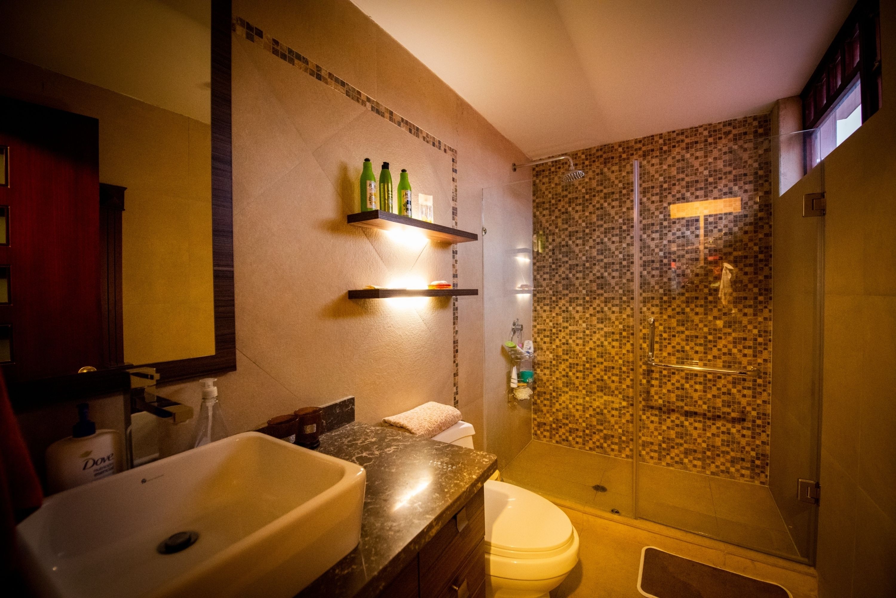A bathroom with a sink, toilet, and shower area. There are shelves with toiletries and a mirror above the sink.