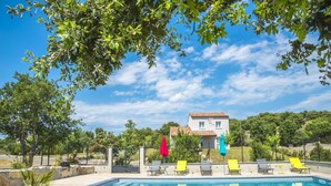 Ein Swimmingpool mit Liegestühlen und Sonnenschirmen, umgeben von viel Grün und einem Haus im Hintergrund.