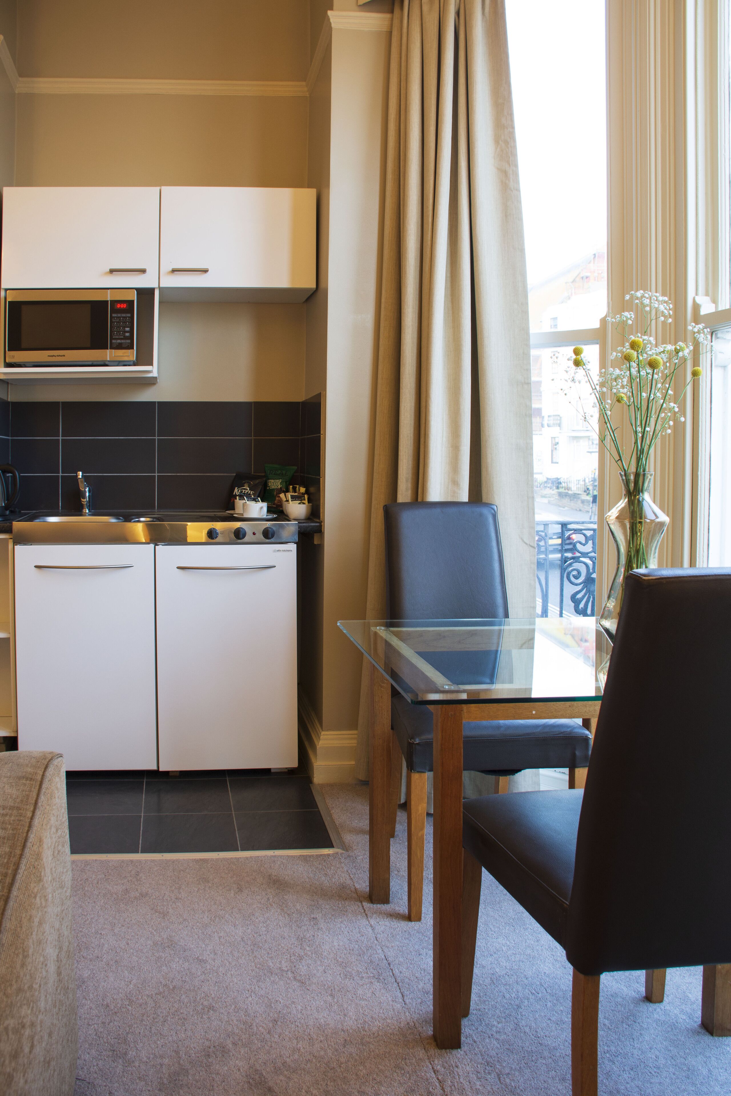 A compact kitchen with a microwave, stove, and cabinets. There is a dining table with chairs and a vase with flowers on the window sill.