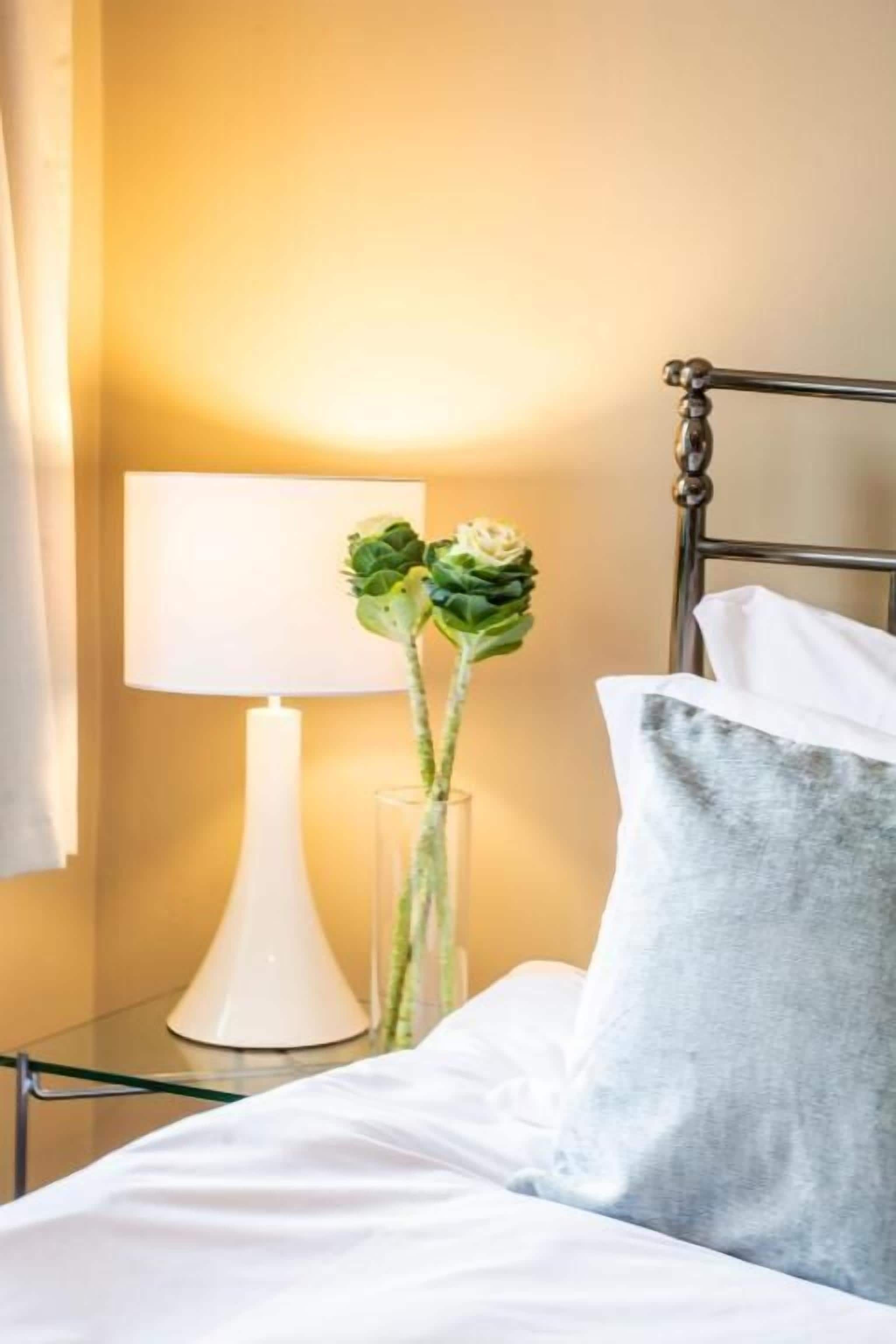 A neatly made bed with white linens, a grey pillow, and a clear vase with green plants on a bedside table.
