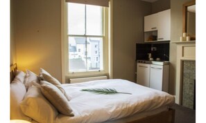 A bedroom with a bed, a window, a kitchen area, and a view of buildings outside.