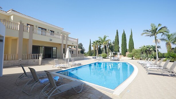 A swimming pool with lounge chairs and umbrellas, surrounded by palm trees and a building with balconies. - Villa Jorja, Fabulous and luxurious 7BDR Villa, Close to Fig tree Bay Beach (Protaras)