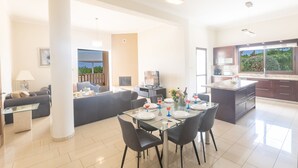 A modern living room with a glass dining table, black chairs, and a sofa. There is a kitchen area with dark cabinets and a large window. - Villa Jorja, Fabulous and luxurious 7BDR Villa, Close to Fig tree Bay Beach (Protaras)