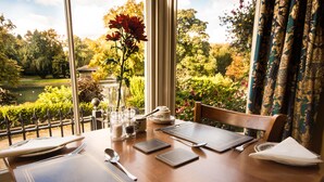 A dining table set with a vase of flowers, a candle, and a view of a garden and lake. - Roseleigh Guest House (Buxton)