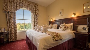 A hotel room with a large bed, floral curtains, a wooden headboard, a bedside table with a radio, and a window with a view of trees. - Roseleigh Guest House (Buxton)