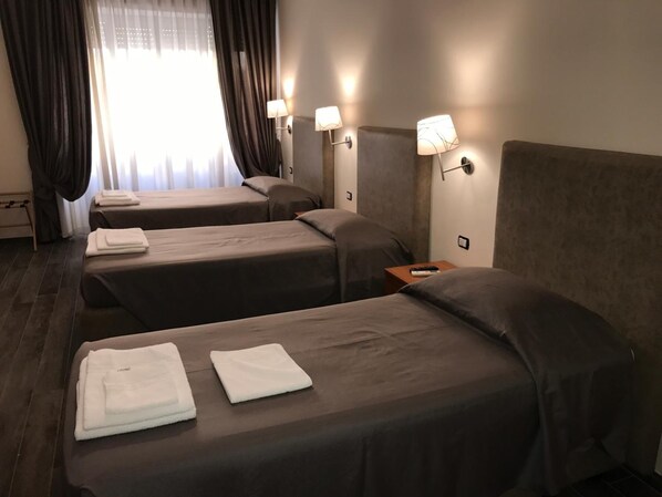 Habitación de hotel con tres camas, cada una con una toalla blanca, una mesita de noche con control remoto y lámparas de pared.