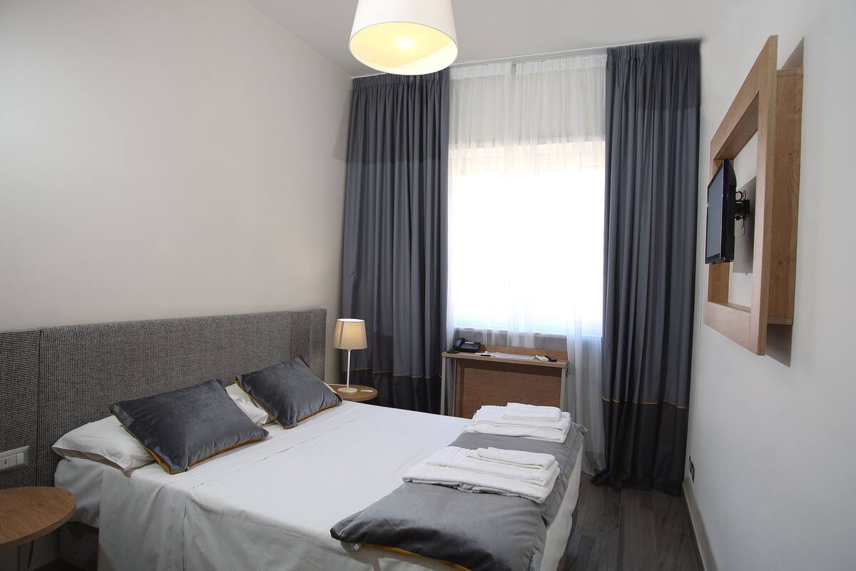 Een hotelkamer met een bed, twee kussens, een nachtkastje met een lamp, een aan de muur bevestigde televisie en een raam met gordijnen.