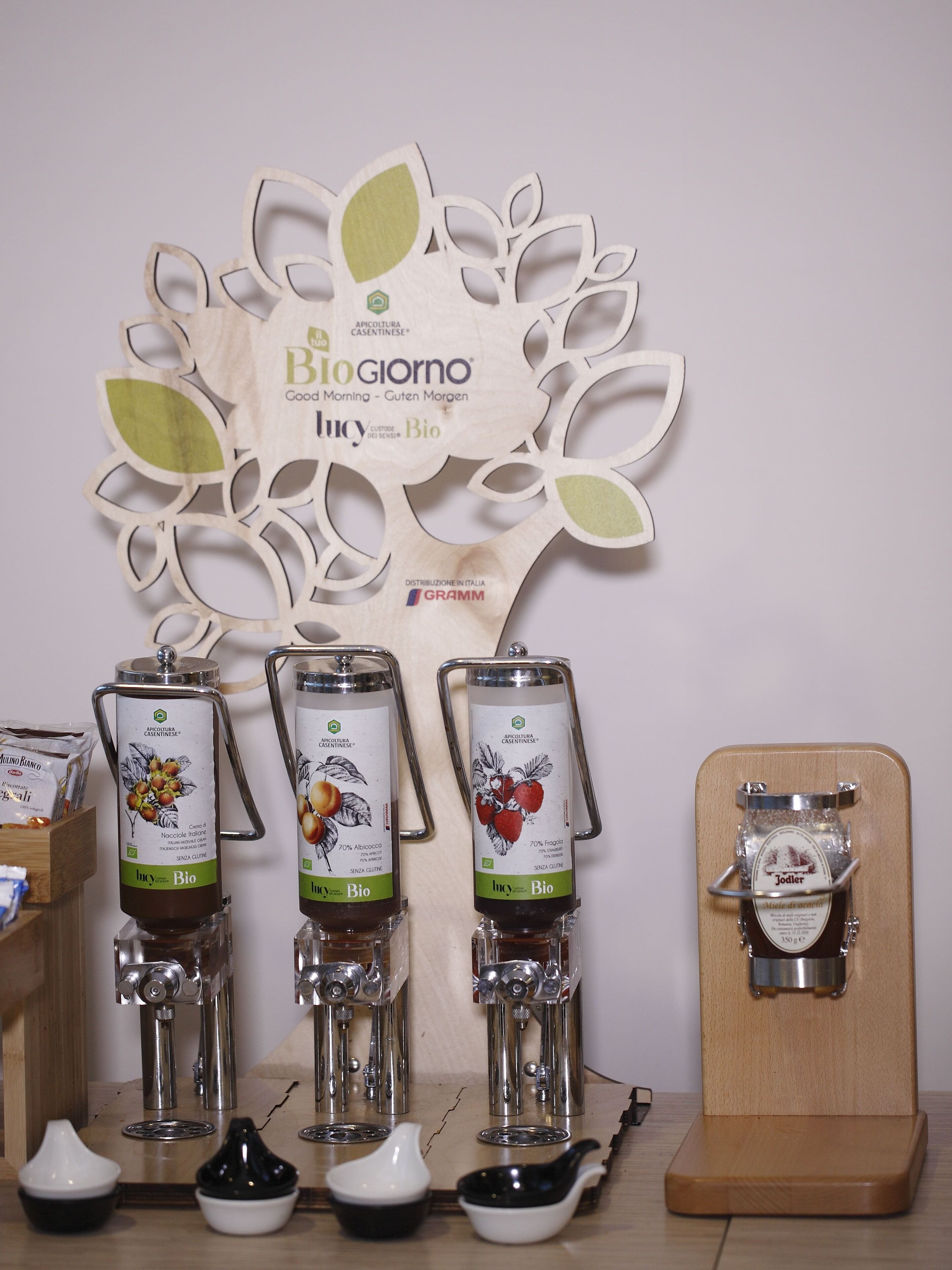 Esposizione di distributori di miele con etichette, un cartello a forma di albero in legno e un ripiano in legno con un vasetto.