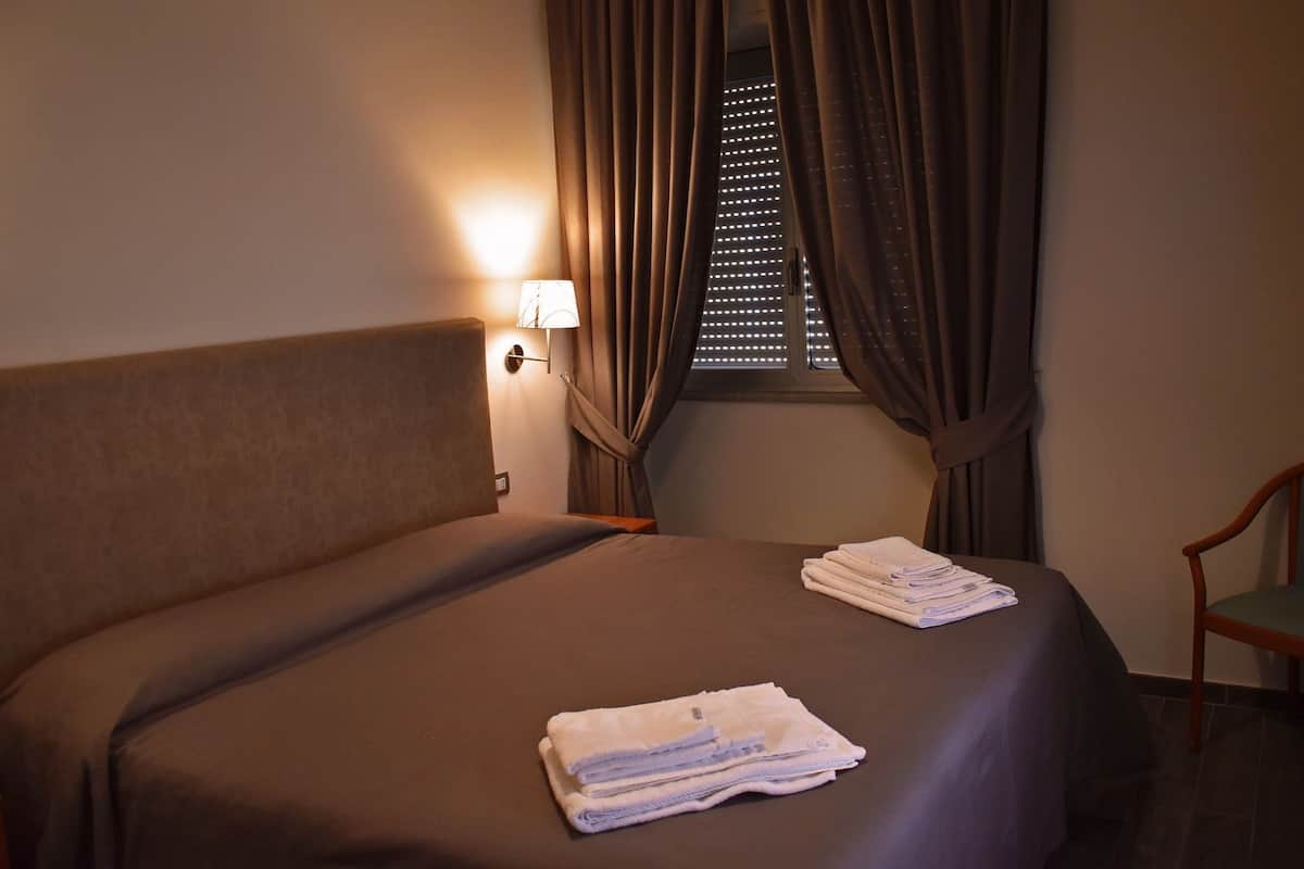 Een hotelkamer met een bed, een nachtkastje, een lamp, een raam met jaloezieën en een stoel.