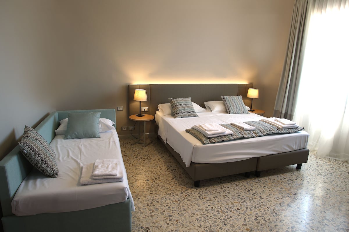 Een hotelkamer met twee bedden, elk met een nachtkastje en lamp, een groot raam met gordijnen, en een gestippelde vloer.