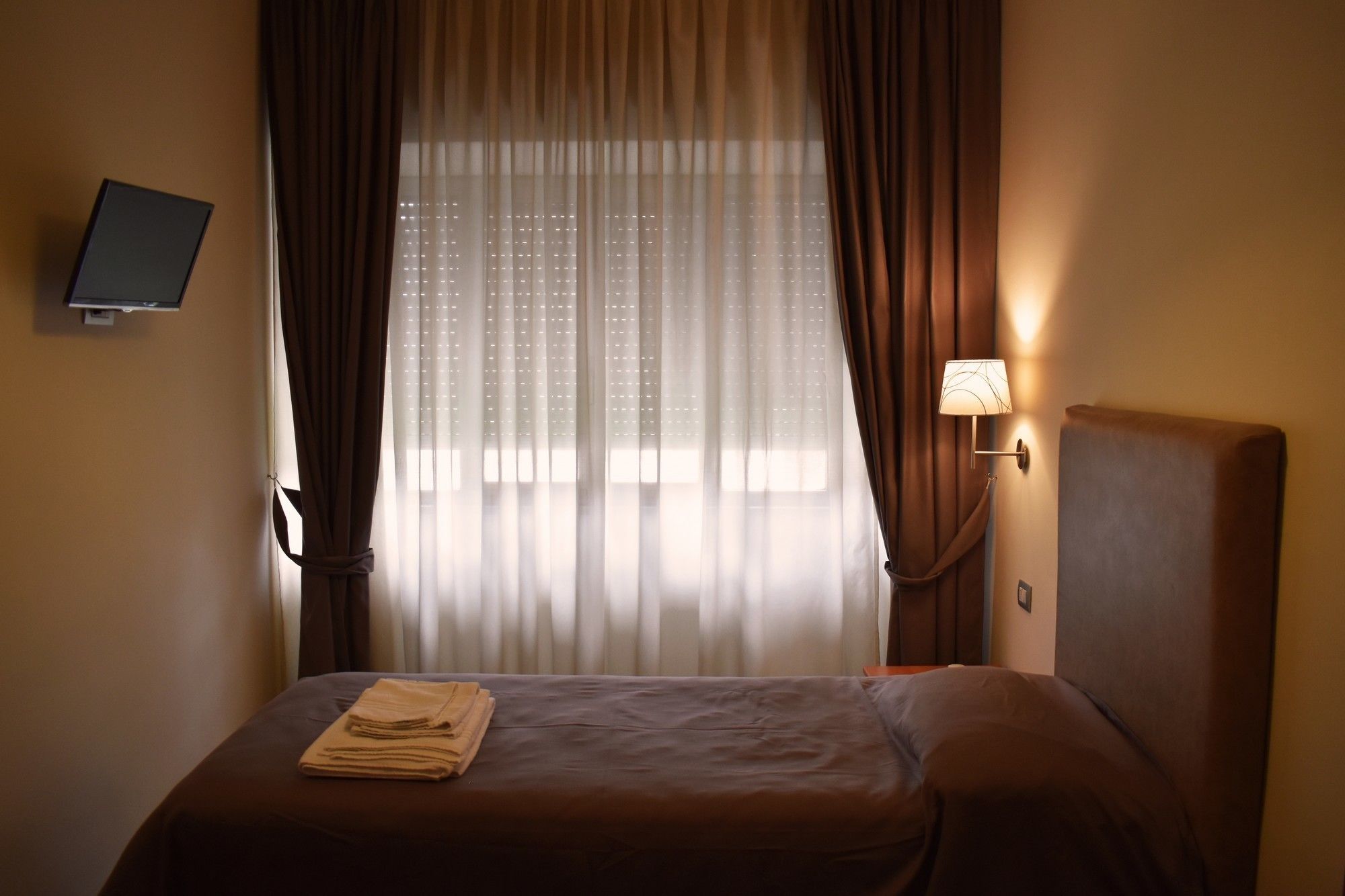 A hotel room with a bed, curtains, a wall-mounted TV, and a bedside lamp.